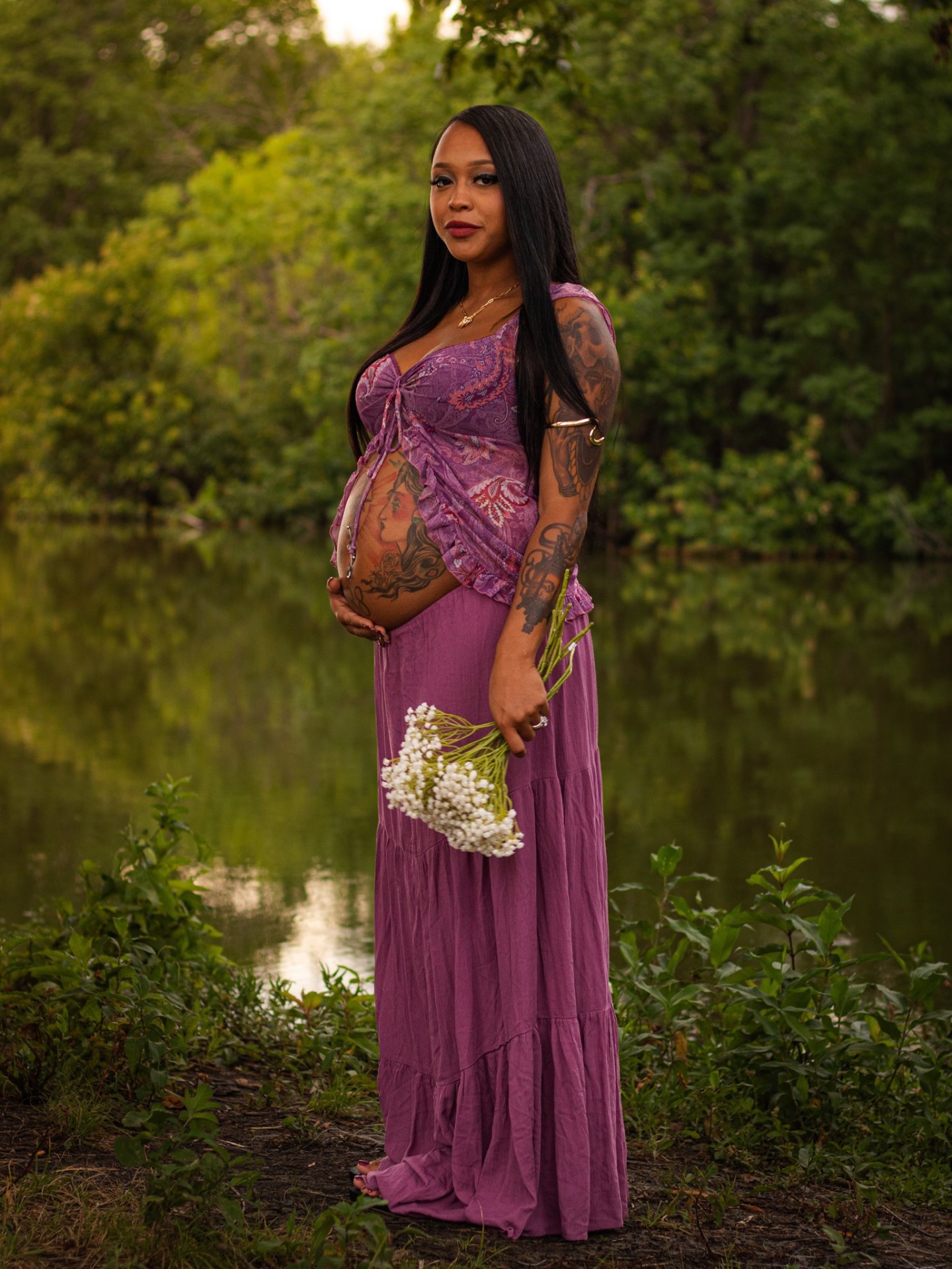 Expecting mother in a purple dress holding baby's breath by a lake at golden hour