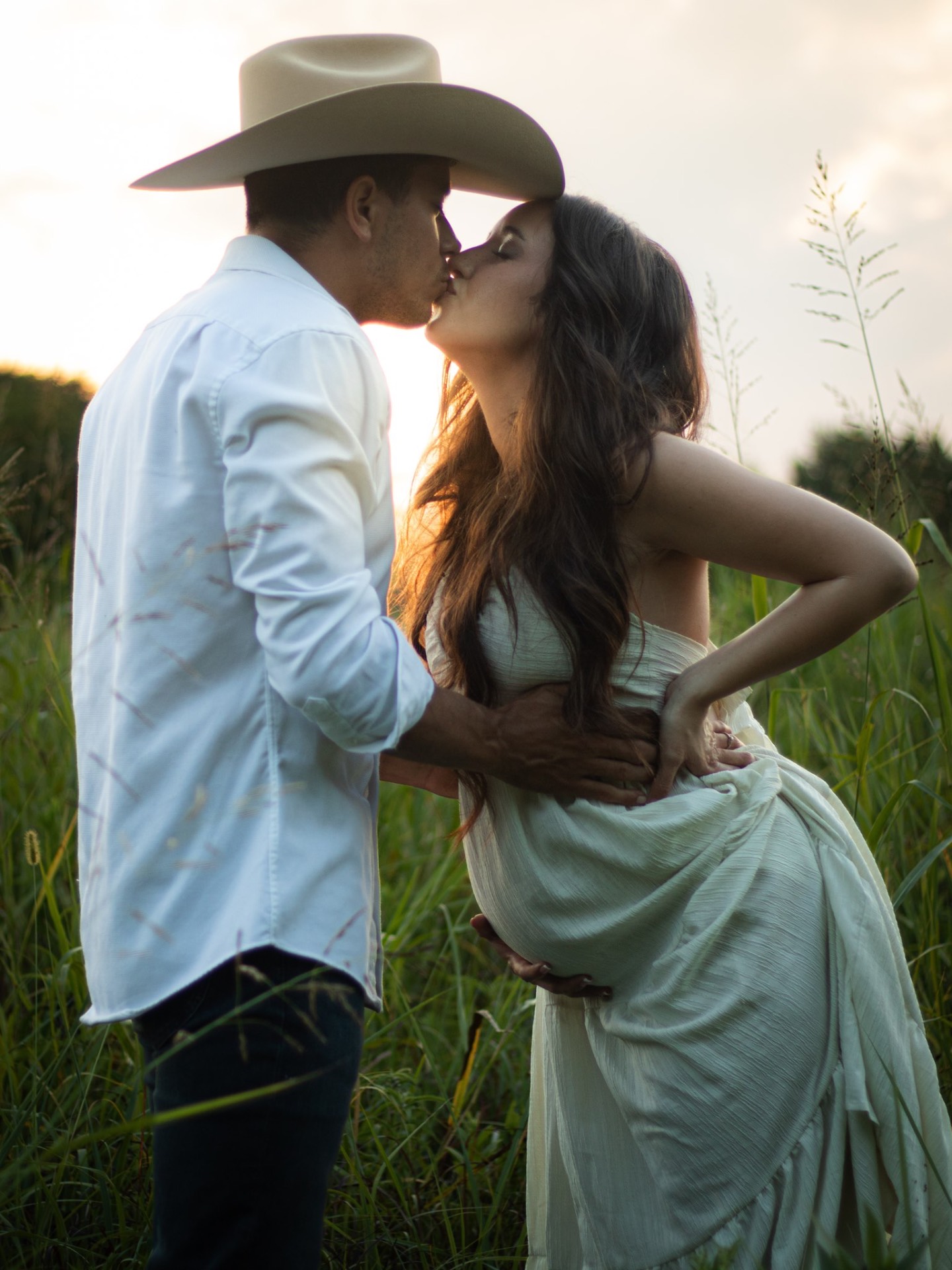 Cowboy kissing his pregnant partner in tall grass at sunset